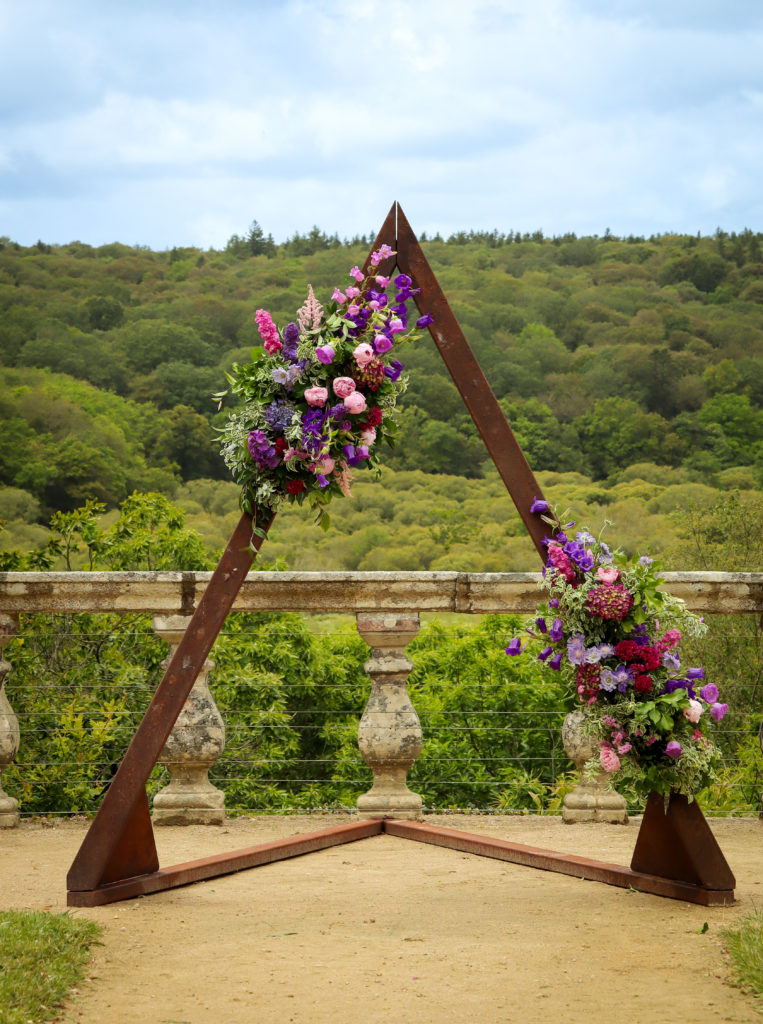 Mariée souriante avec bouquet de fleurs écoresponsables devant arche florale, fleuriste mariage Guidel Bretagne.